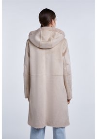 Žena viděná zezadu, oblečená v béžovém kabátu z ovčí vlny s kapucí a světlých modrých džínách na jednobarevném světlém pozadí.