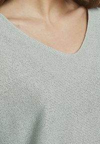 Hellgrüner Strickoberteil mit V-Ausschnitt, strukturiertem Stoff und lockerer Passform. Das Garn zeigt ein feines, offenes Webmuster.