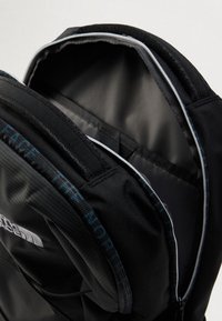 Czarny plecak z wzorzystą powierzchnią, ciemnymi zamkami błyskawicznymi oraz szarym wyściełaniem wnętrza. Projekt obejmuje wyściełane przegrody i detal z logo.