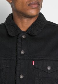 Černá džínová bunda s texturovaným černým sherpa límcem, stříbrnými knoflíky a viditelným červeným štítkem Levi's na levé kapse.