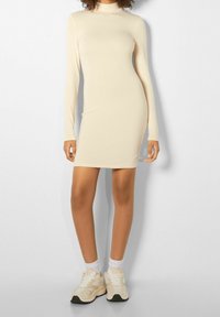 Ihonmyötäinen, pitkähiha beige mekko, jossa korkea kaulus ja sivuhalkiot, yhdistetty valkoisiin sukkiin ja beigen sävyisiin lenkkareihin. Sileä, joustava materiaali.