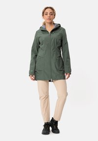 Giacca impermeabile verde con cappuccio, zip frontale, due tasche laterali e polsini elasticizzati, abbinata a pantaloni beige e scarponi da trekking neri.