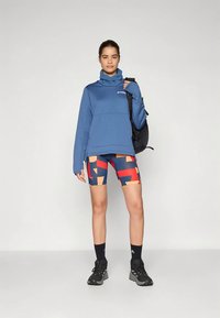 Egy nő kék, magas nyakú pulóverben, geometrikus mintájú sortban, fekete zokniban és tornacipőben, fekete hátizsákot tart a vállán.