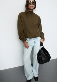 Olive zielona kurtka zapinana na zamek, luźny krój, z wysokim kołnierzem i bocznymi kieszeniami; jasnoniebieskie dzwony; czarna skórzana torebka; duże okulary przeciwsłoneczne.