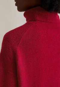 Roter Wollpullover mit Rollkragen und strukturiertem Finish. Verfügt über einen hohen Kragen und Raglanärmel und zeigt eine satte Farbe mit subtilen Variationen.