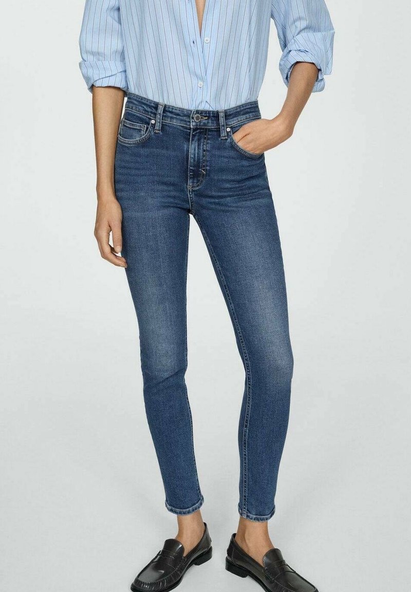 Jean skinny bleu foncé taille haute, avec cinq poches et un léger délavage. Associé à une chemise boutonnée bleu clair à rayures.