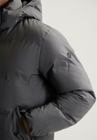 Pufferjack in donkergrijs met een matte afwerking, voorzien van een capuchon, zijzakken en een reliëflogo op de mouw.