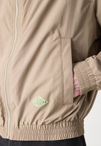 Beige lett jakke med full glidelås, elastiske ermer og et grønt gummimerke. Har to sidelommer og glatt tekstur.