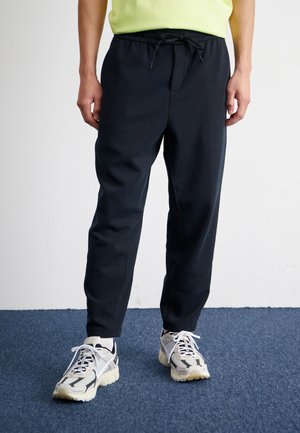 Schwarze Jogginghosen mit elastischem Bund und Kordelzug, ausgestattet mit Seitentaschen und einem taillierten Beinprofil. Kombiniert mit Sneakers in neutralen Farbtönen.