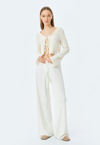 Witte cropped sweater met textuurbreiwerk, gestrikte sluiting aan de voorkant en uitlopende mouwen, gecombineerd met brede witte textuurbroeken.