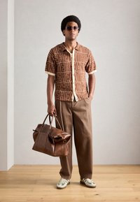 Short-sleeved brown shirt with large cream letters, beige collar; brown wide-leg pants; brown leather duffel bag; green and white sneakers.