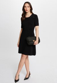 Zwart kortemouwjurk met een getailleerde bovenkant en een uitlopende rok. Heeft decoratieve knopen op de schouder. Gecombineerd met een textuur zwarte clutch.