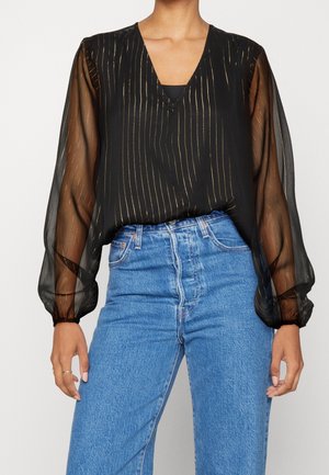 Kobieta w czarnej bluzce z dekoltem w kształcie litery V, ozdobionej złotymi pionowymi paskami i przezroczystymi długimi rękawami, w połączeniu z niebieskimi dżinsami z wysokim stanem.