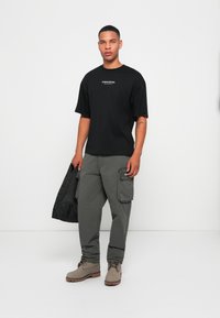 Man met een zwart "Studio Edition" t-shirt, een grijze cargobroek, beige laarzen, die een zwart gewatteerd jack vasthoudt, staande tegen een witte achtergrond.