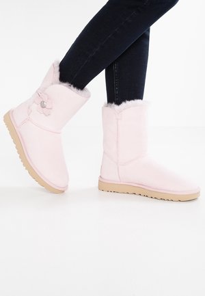 Fødder iført lyserøde midt-på-læret støvler med pelsfor og blomsterdetalje, kombineret med mørke, tætsiddende jeans mod en hvid baggrund.