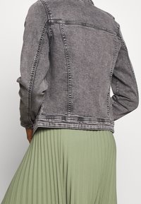 Grå denimjacka med en figursydd design, synliga sömdetaljer och långa ärmar. I kombination med en ljusgrön plisserad kjol.