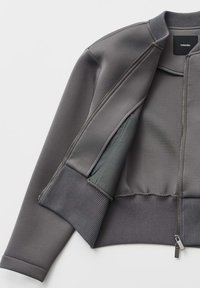 Giacca bomber grigia con chiusura lampo, polsini e orlo a coste, tessuto liscio e tasca interna con fodera a contrasto.