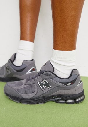 Jambes portant des chaussettes blanches et des baskets grises New Balance 2002R, debout sur un sol vert contre un mur blanc.