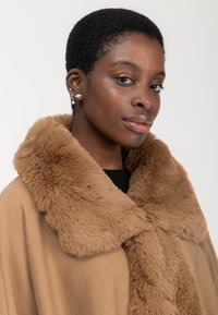 Giovane con capelli corti e ricci, indossa più orecchini e un cappotto color cammello con un ampio colletto morbido, guardando con sicurezza verso la fotocamera.