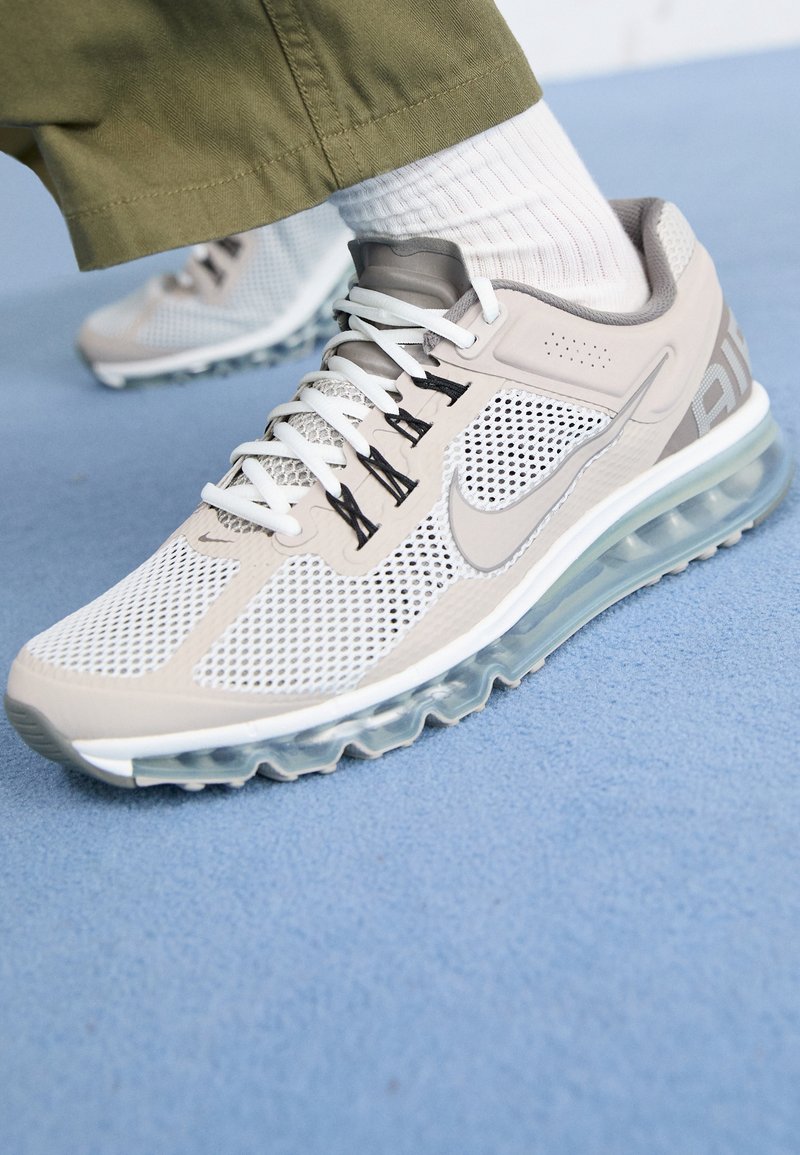 Chaussure de course Nike beige clair avec lacets blancs, empiècements en mesh et semelle à coussin d’air visible sur une personne portant des chaussettes blanches et un pantalon olive.