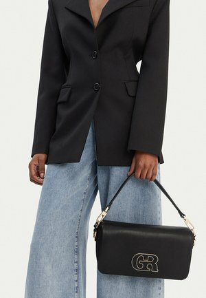 Persona vistiendo un blazer negro y jeans de pierna ancha azul claro, sosteniendo un bolso negro con el logo dorado "GR" y asa.