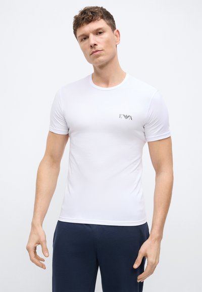 Νεαρός άνδρας με κοντά κατσαρά μαλλιά, φορώντας ένα εφαρμοστό λευκό μπλουζάκι με μικρό μαύρο λογότυπο στο στήθος και σκουρό μπλε παντελόνι.