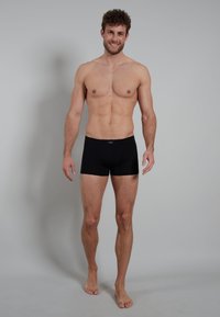 Shorts de boxe noirs en tissu lisse, dotés d'une taille confortable et d'une coupe ajustée. Le modèle se tient pieds nus sur un fond clair.