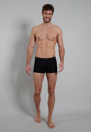 Zwarte boxershorts van soepel materiaal, met een comfortabele tailleband en een aansluitende pasvorm. Model staat blootsvoets tegen een lichte achtergrond.
