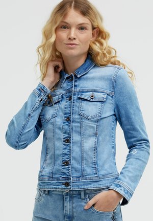 Junge Frau mit blonden lockigen Haaren, die eine hellblaue Jeansjacke und passende Jeans trägt, eine Hand berührt den Kragen, die andere in der Tasche.