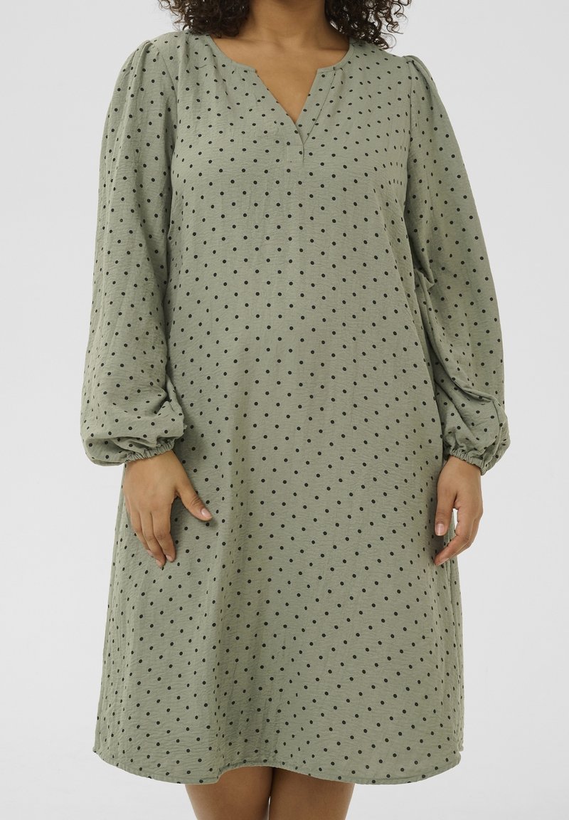 Femme portant une robe ample vert clair à pois noirs avec de longues manches bouffantes, debout sur un fond uni.