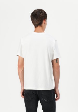 Hombre con cabello corto y castaño de espaldas, que lleva una camisa blanca sencilla de manga corta y jeans negros con un pequeño tatuaje en el brazo izquierdo.