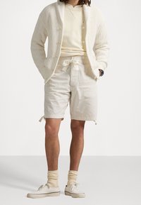 Muž oblečený v krémovém pleteném cardiganu, světle šedé mikině, béžových kraťasech s šnůrkou, krémových ponožkách a bílých plátěných teniskách, stojící s rukama v kapsách.