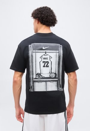 Mann mit lockigem Haar, der ein schwarzes T-Shirt trägt, zeigt eine Grafik eines Basketballtrikots, von Schuhen und einem Ball im Spind auf dem Rücken.