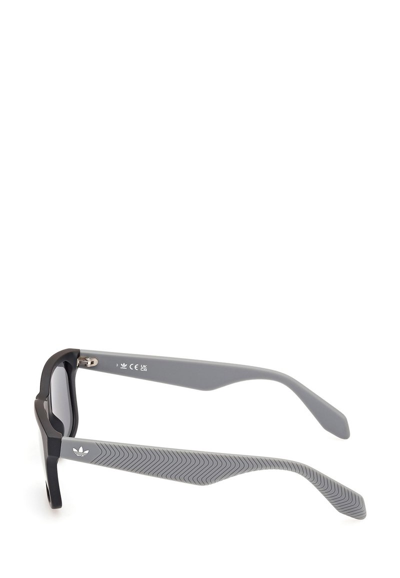 Black sunglasses with a thick frame and gray arms featuring a textured pattern. Lenses are tinted gray, with a prominent logo on the side.