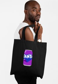 Zwarte tote bag met een paarse afbeelding van een Fanta druivensoda blikje. Het ontwerp bevat vetgedrukte witte letters op een gladde textuur.