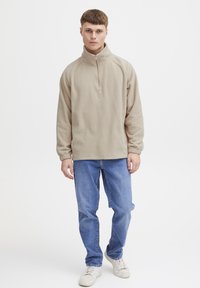 Beige fleece pullover with a high collar and half-zip, featuring ribbed cuffs and an embroidered emblem on the chest. Paired with blue jeans.