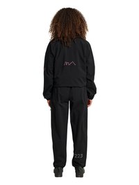 Person with curly hair wearing a black jacket and pants with a pink skyline design on the jacket back and "223" on the right pant leg.