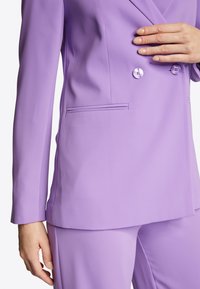 Liliowy, dopasowany żakiet o gładkiej fakturze, z podwójnym zapięciem na guziki i boczną kieszenią. Długie rękawy mają schludny, wyrazisty krój.