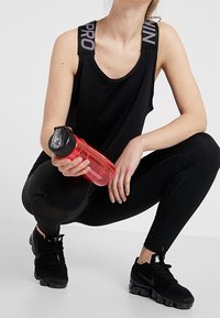 Černý tílko s nápisem "PRO" na ramínkách, černé legíny, černé tenisky, držící průhlednou červenou láhev na vodu, v pokrčené pozici.