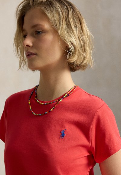 Rövid szőke hajú fiatal nő piros pólót visel kék hímzett logóval és színes gyöngysorokkal, balra néz.