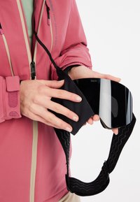 Skibrille mit schwarzem Glas, flexiblem Band mit wellenförmiger Textur und einem schwarzen Mikrofasertuch, das von einer Person in einer pinken Jacke gehalten wird.