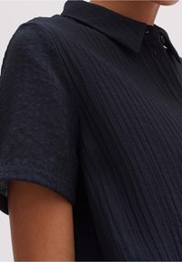 Marinblå kortärmad skjorta med en strukturerad, ribbad mönstring, en klassisk krage och en enda knap på halsen. Mjukt tyg.