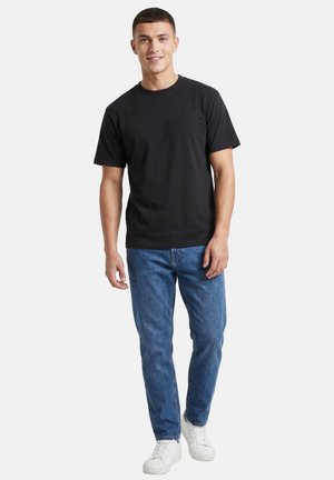 Ung mand står iført en ensfarvet sort t-shirt, blå jeans og hvide sneakers mod en hvid baggrund.