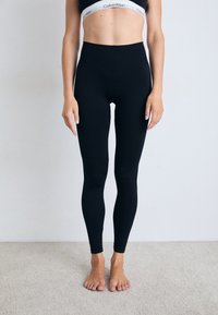 Donna che indossa leggings neri a vita alta e un reggiseno sportivo nero di Calvin Klein, in piedi a piedi nudi su un tappeto chiaro contro un muro semplice.