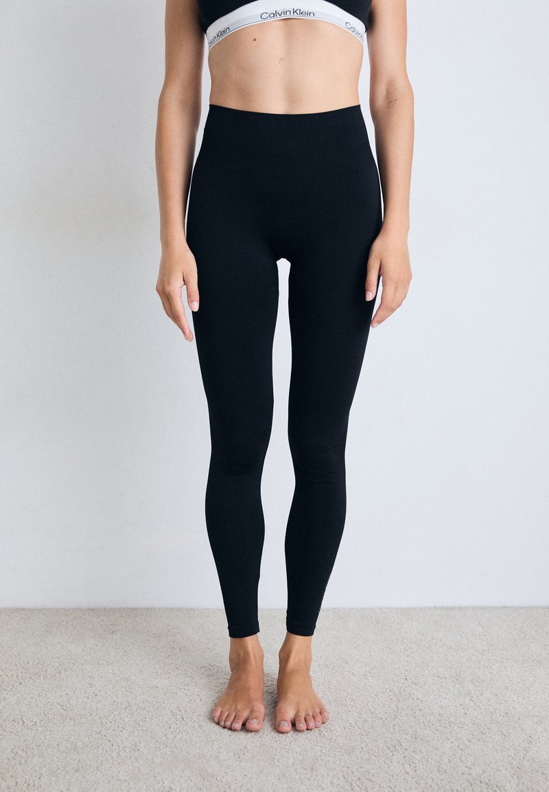Donna che indossa leggings neri a vita alta e un reggiseno sportivo nero di Calvin Klein, in piedi a piedi nudi su un tappeto chiaro contro un muro semplice.