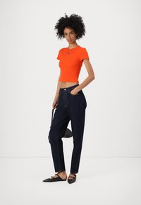 Lyhythihainen oranssi crop top, joka on yhdistetty tummansinihin korkeavyötäröisiin farkkuihin. Mallilla on päällään mustat tasapohjaiset kengät ja hän pitää käsissään pientä mustaa laukku.