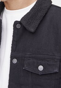 Veste en jean noire avec col en shearling, deux poches poitrine, boutons-pression argentés et tissu texturé. Chemise blanche visible en dessous.