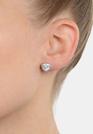 Pendiente de stud en forma de corazón con una gran piedra preciosa clara en un engaste de metal tono plateado, posicionado contra un primer plano de una oreja.