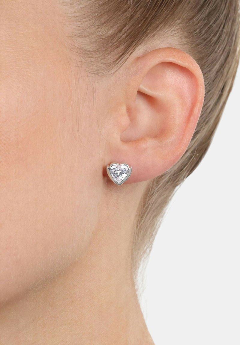 Hjärtförformad stiftörhänge med en stor klar ädelsten i en silverfärgad metallram, placerad mot en närbild av ett öra.