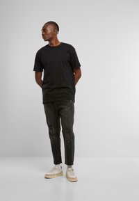 Hombre con cabello trenzado que lleva una camiseta negra, jeans negros desgastados con manchas de pintura, zapatillas blancas, de pie con las manos detrás de la espalda.